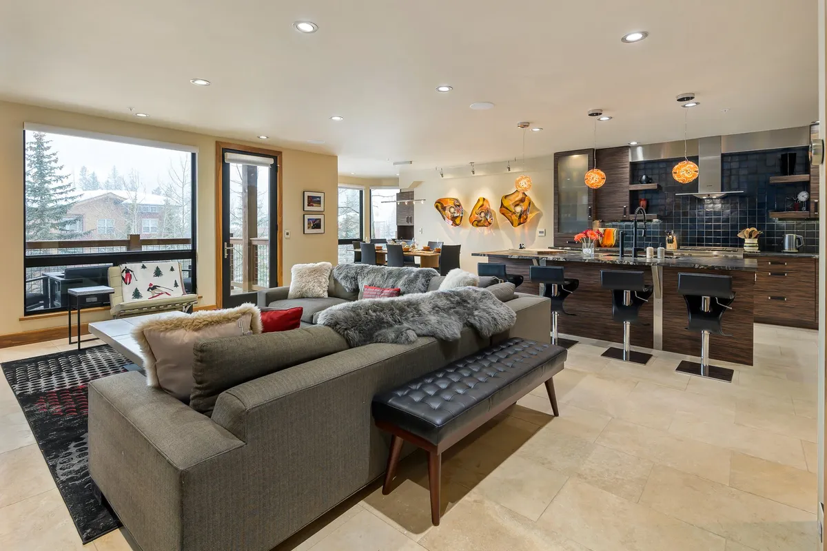 This image displays an open-concept living area where the kitchen, featuring a large island, flows seamlessly into a dining and lounging space with a large sectional sofa and artistic touches. Bright natural light pours in through large windows, creating a welcoming environment for gatherings.