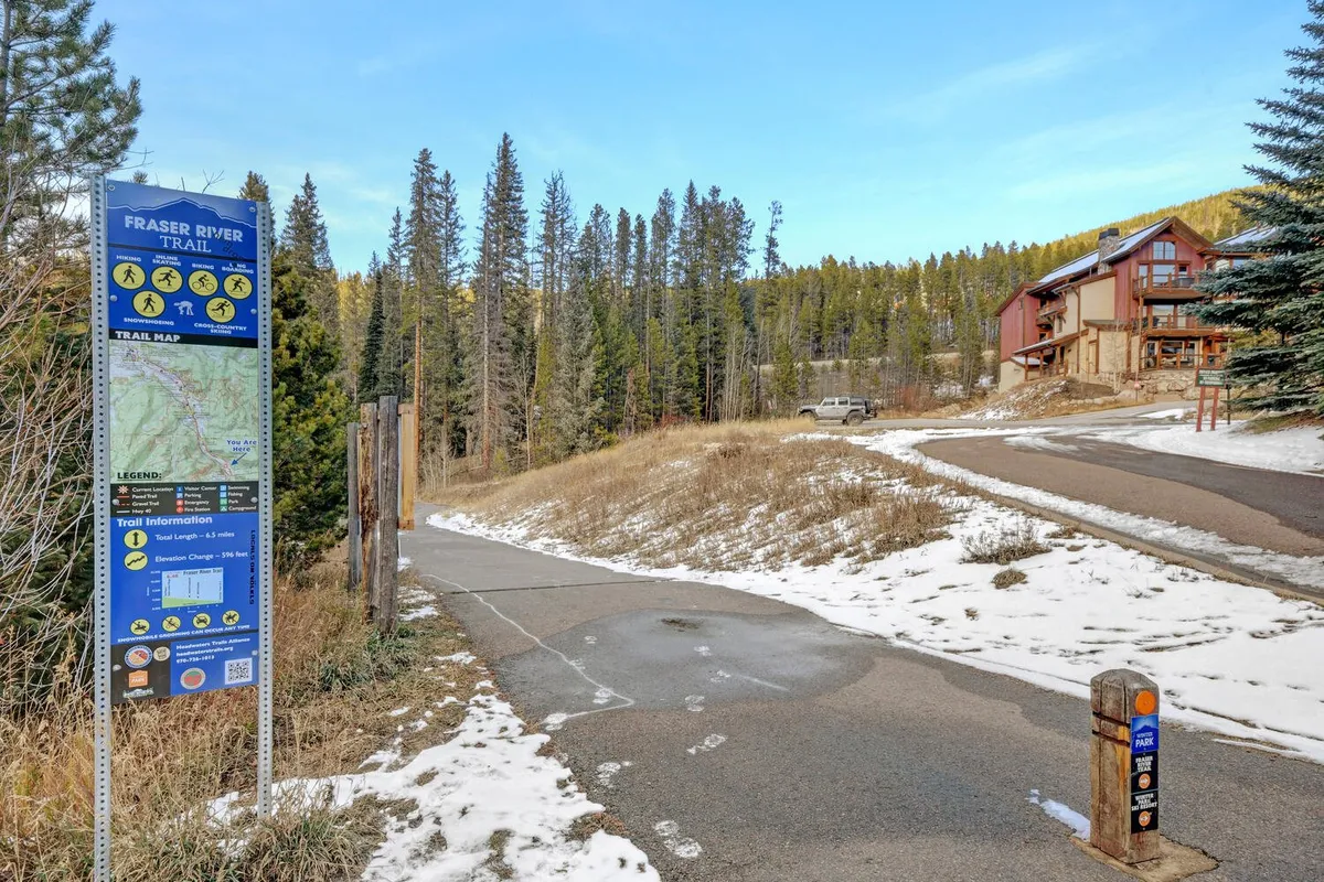 A paved trail winds through snow-dusted trees near a residential building, marked by a trail sign. The environment is crisp and inviting, ideal for hiking, biking, and nature walks along the Fraser River Trail or to nearby adventure spots.