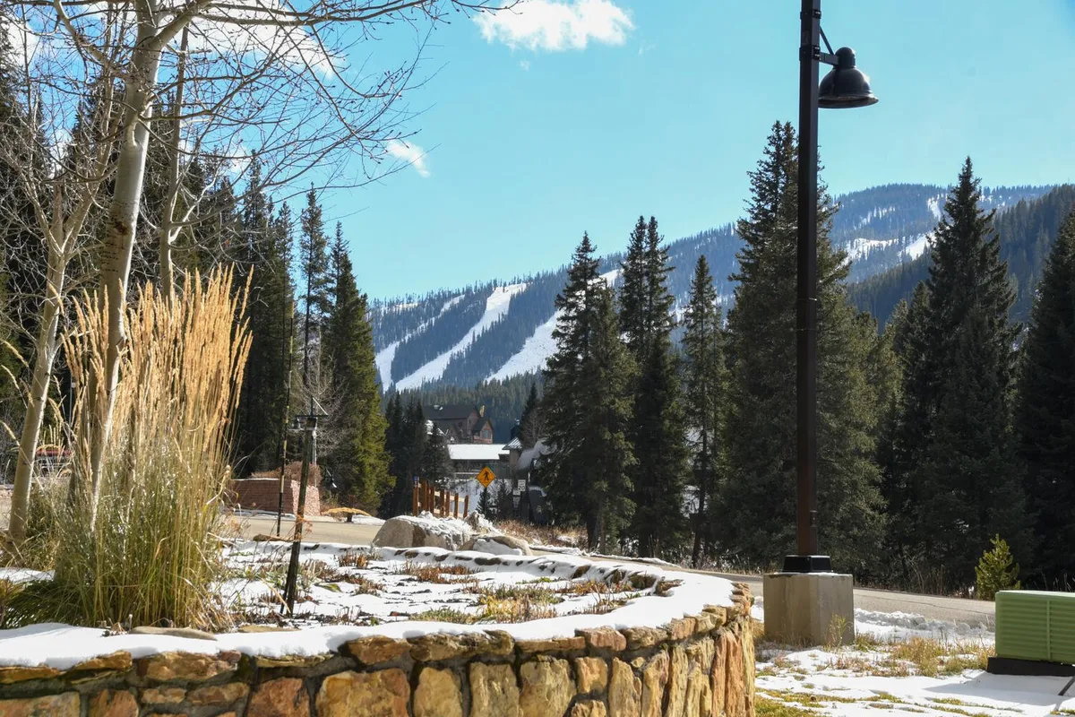 A tranquil winter landscape with snow-covered mountains, tall evergreen trees, decorative landscaping, and a nearby road sign. The image evokes peace and proximity to nature, highlighting the scenic location and accessibility of the condo to outdoor adventures.