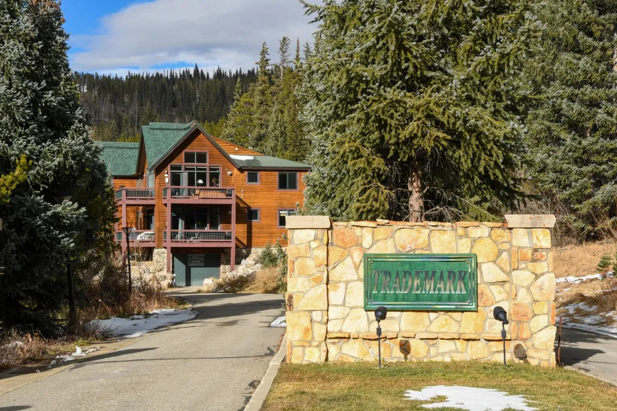 A picturesque view of a wooden cabin nestled among mature trees, with a stone entrance sign and a driveway leading up to the house. The welcoming porch offers space to relax and enjoy the natural surroundings, while the overall setting is peaceful and private—suggesting the perfect place for couples or small groups to unwind in nature.