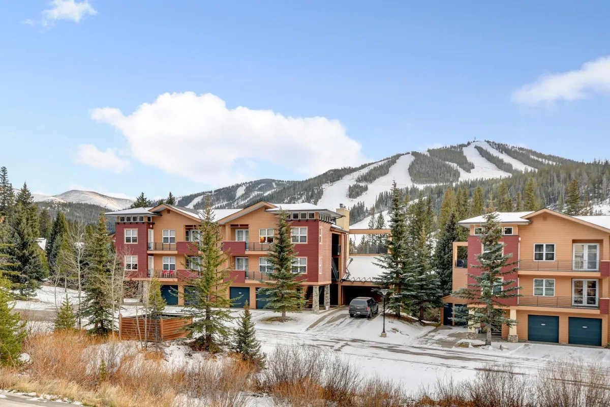 A striking exterior view showcases snow-covered mountains behind colorful building exteriors and tall pine trees. The scene is vibrant with the sense of adventure and the promise of outdoor activities like skiing, snowboarding, hiking, and nature walks.