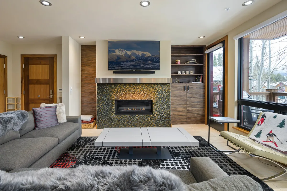 A contemporary great room featuring a modern fireplace, large windows that fill the space with natural light, eye-catching artwork, and a unique coffee table. The inviting space is arranged for relaxation and entertaining, with views outside that hint at the Rocky Mountain scenery beyond.