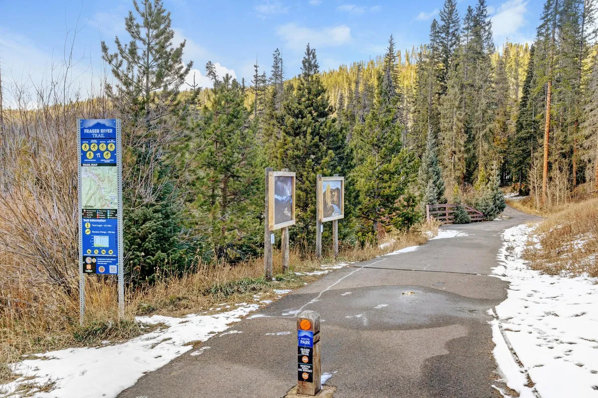 A lush, tree-lined pathway marked by clear trail signs, with a smooth walking and biking path leading into the woods. The scene evokes the sense of discovery, fresh mountain air, and connection to nature, ideal for family strolls or bike rides along the Fraser River Trail.