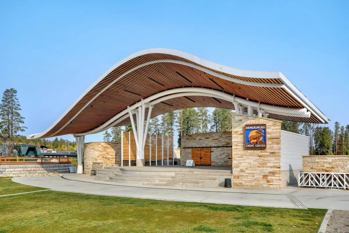 An exterior shot of a distinctive amphitheater with a curved roof and stone architecture, set outdoors with steps leading up to the stage. The space is primed for live music, concerts, and outdoor gatherings under open skies, evoking a festive, communal atmosphere.