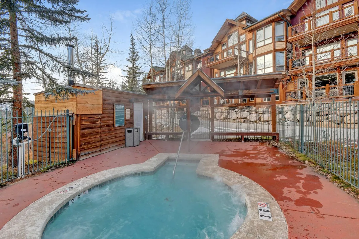 A tranquil outdoor hot tub sits beneath a wooden structure, surrounded by trees and the exterior of the building. The scene evokes relaxation and camaraderie after a day of adventure, with nature as the backdrop.