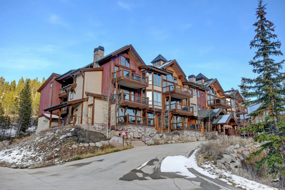 A wide-angle exterior view of a multi-level mountain lodge with wooden balconies, surrounded by mature trees and stone pathways. The scene suggests both relaxation and access to outdoor amenities, including spaces ideal for barbecues and fire pit gatherings.