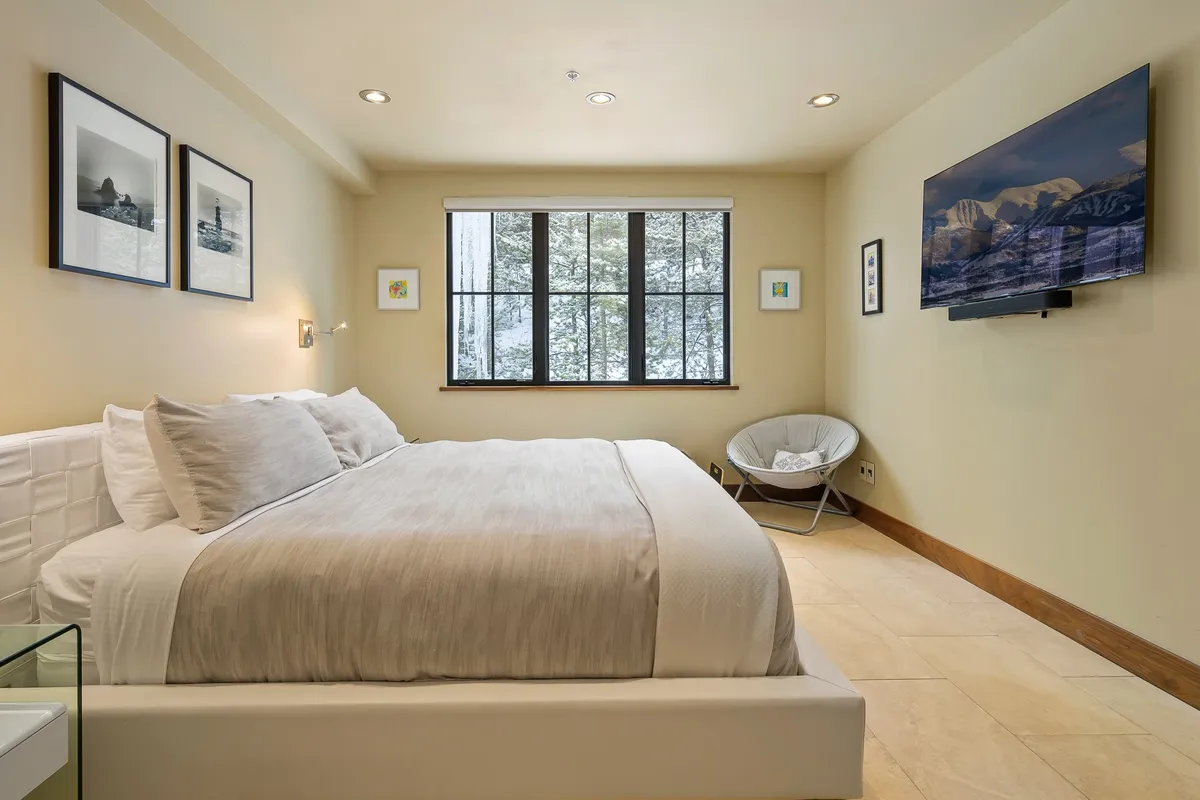 A serene bedroom featuring a plush bed, a large window allowing natural light to fill the room, a wall-mounted television, and tasteful artwork on the walls. The space is designed for relaxation and comfort, with a calming color palette and soft bedding.