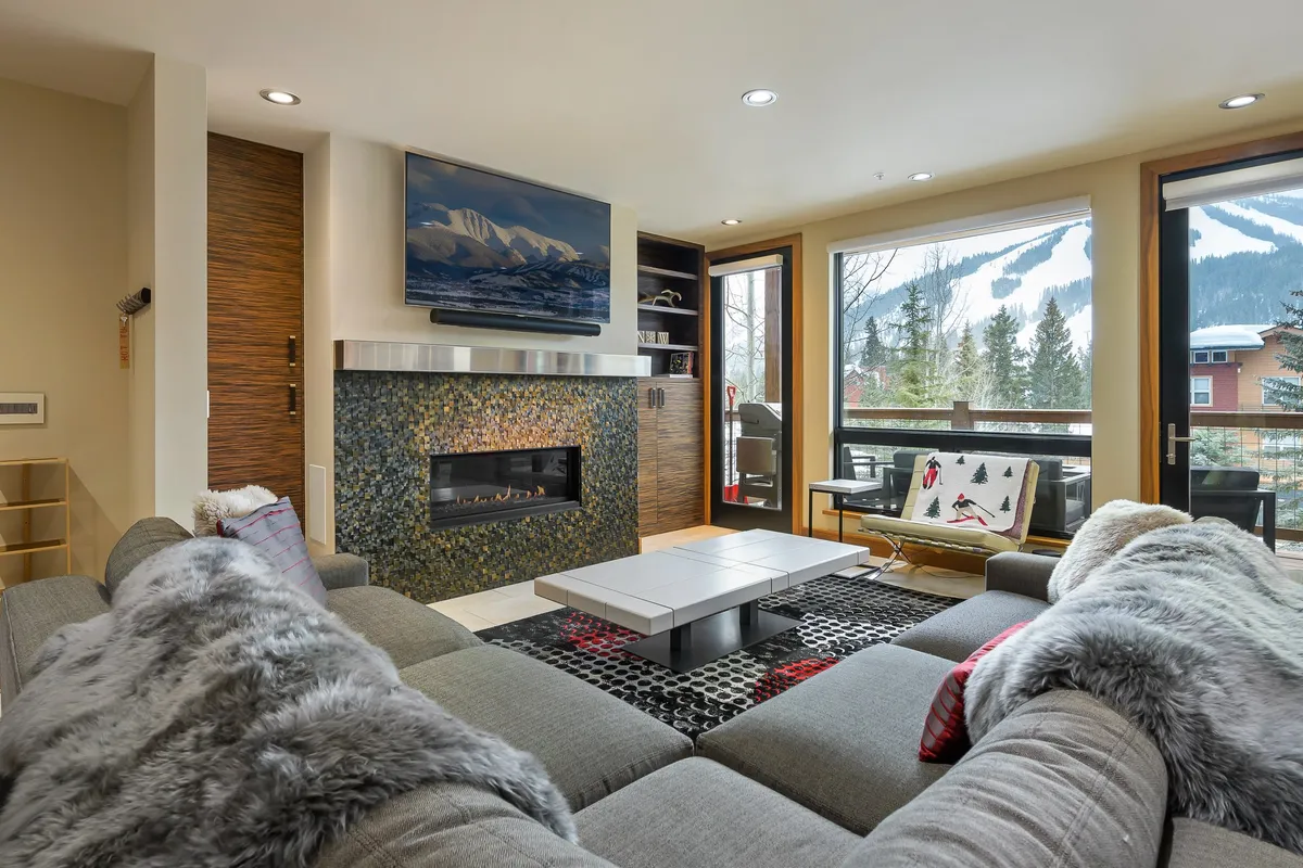 A spacious living room featuring a crackling fireplace, plush sofa seating, and expansive windows framing breathtaking mountain views. Snow gently falls outside, while the warm, inviting interior offers the perfect space for relaxation or gathering with friends and family after a day on the slopes.
