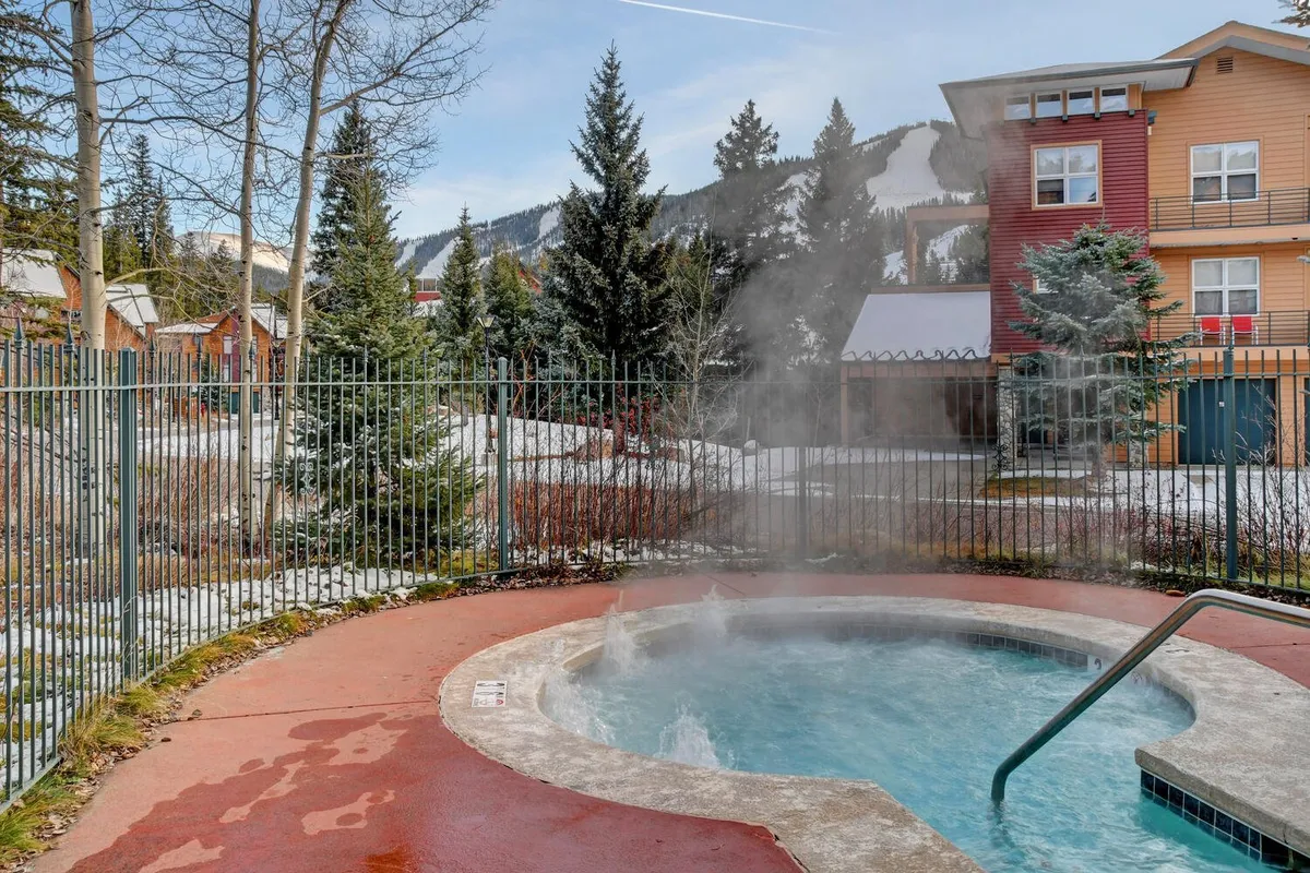 This exterior shot highlights a steaming hot tub nestled among snow-covered trees, adjacent to nearby buildings. The inviting water, surrounded by nature, suggests a place to unwind after a day on the slopes, while the snow-laden landscape emphasizes the mountain setting.